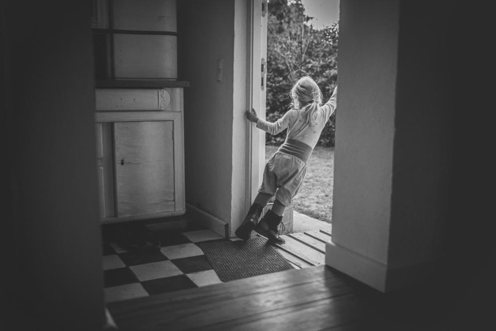 Kleines Mädchen mit Zopf auf Zehenspitzen schaut aus der Gartentür in Mamas Stiefeln
