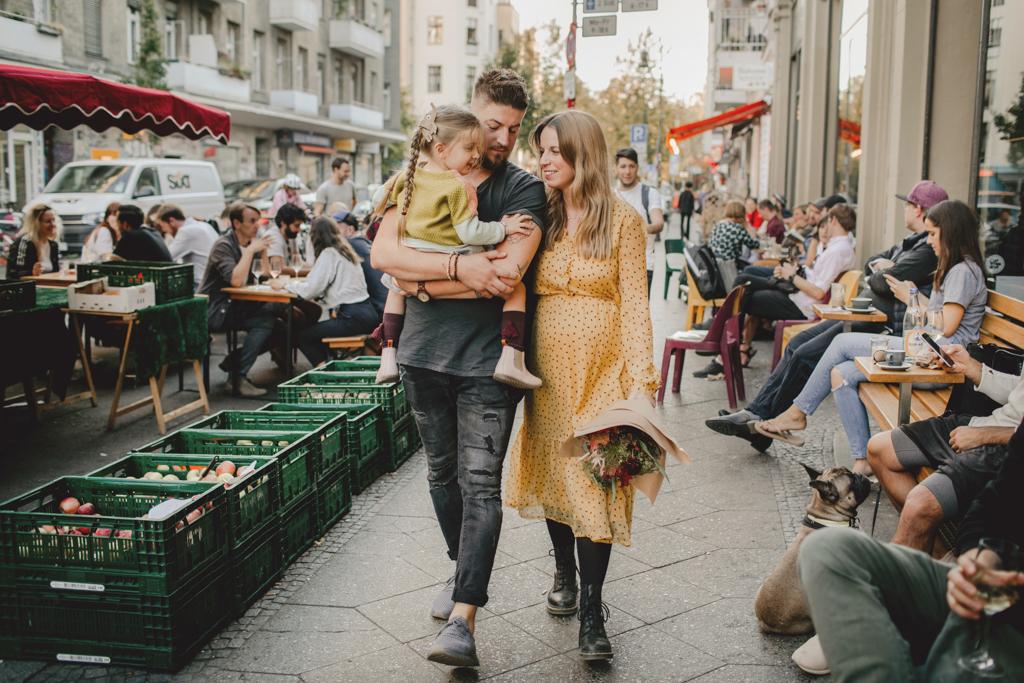 Schwangere Frau mit Babybauch und Blumenstrauß, begleitet von ihrem Mann mit Tochter auf dem Arm, spazieren über einen Markt