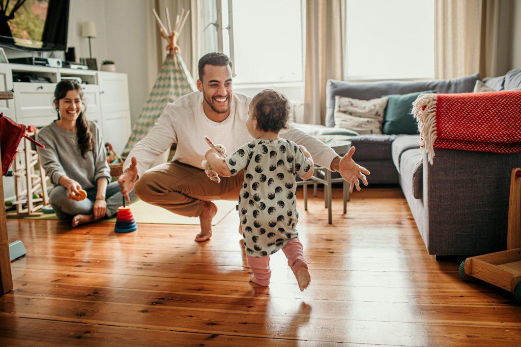 Familie sitzt im Zimmer, Kind rennt auf Papa zu