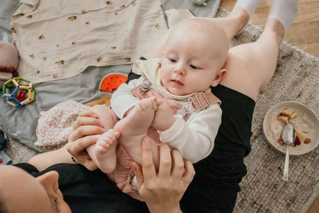 Babyfotograf - Ein Baby liegt im Scho&szlig; der Mama. Sie h&auml;lt liebevoll beim Fr&uuml;hst&uuml;ck die F&uuml;&szlig;e des Babys. Das Baby schaut neugierig zu.