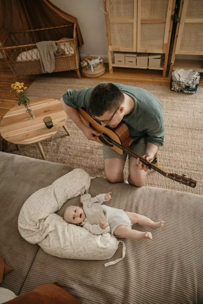 Baby liegt im Stillkissen auf dem Sofa, w&auml;hrend der Papa Gitarre spielt