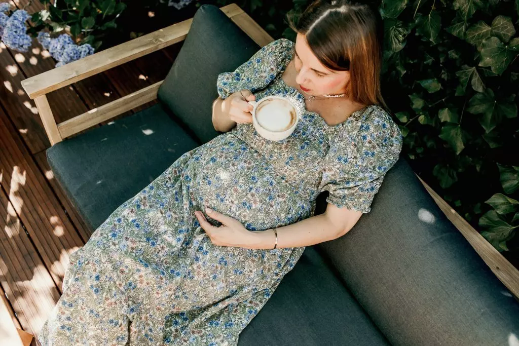 Schwangerschaftsfotografie - Eine schwangere Frau sitzt auf einer Gartenbank im Schatten und genie&szlig;t entspannt ihren Cappuccino.