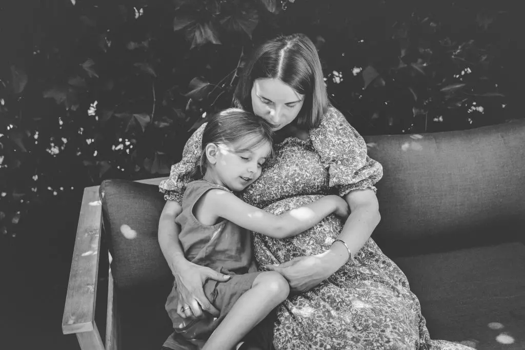 Babybauchfotos bei Schwangerschaftsfotografie in Berlin - Ein M&auml;dchen kuschelt liebevoll ihre Mama mit Babybauch im Garten.