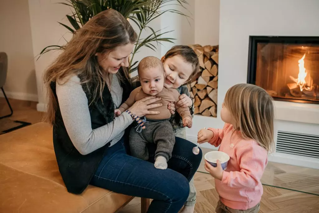 Familienreportage mit Baby - Eine gl&uuml;ckliche Mama h&auml;lt ihr Baby vor dem Kamin, w&auml;hrend ihre Kinder das Baby kuscheln und neugierig anschauen.