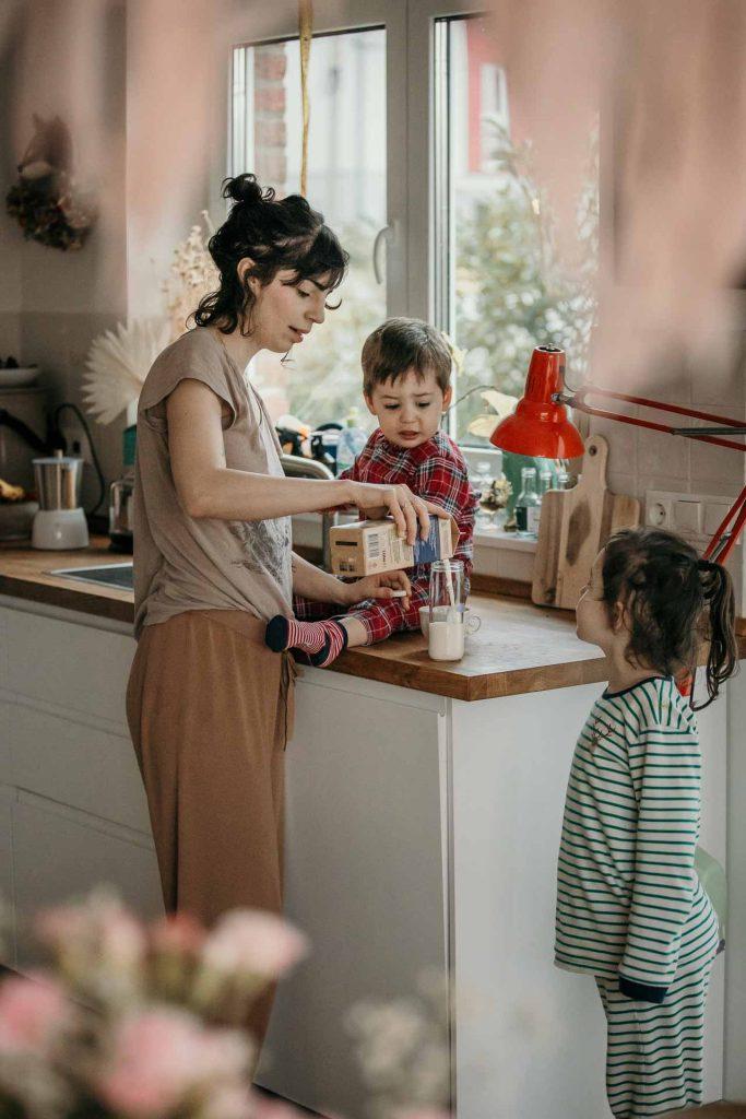 Familienfotos Berlin Brandenburg, Sohn im Schlafanzug auf der Küchenarbeitsplatte, Tochter schaut zu, Mama gießt ein