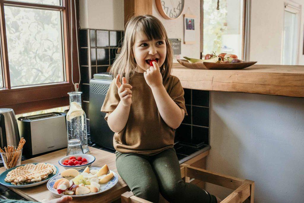 Familienfotoshooting zu Hause Berlin, große Tochter sitzt auf der Küchenarbeitsplatte und nascht eine Himbeere