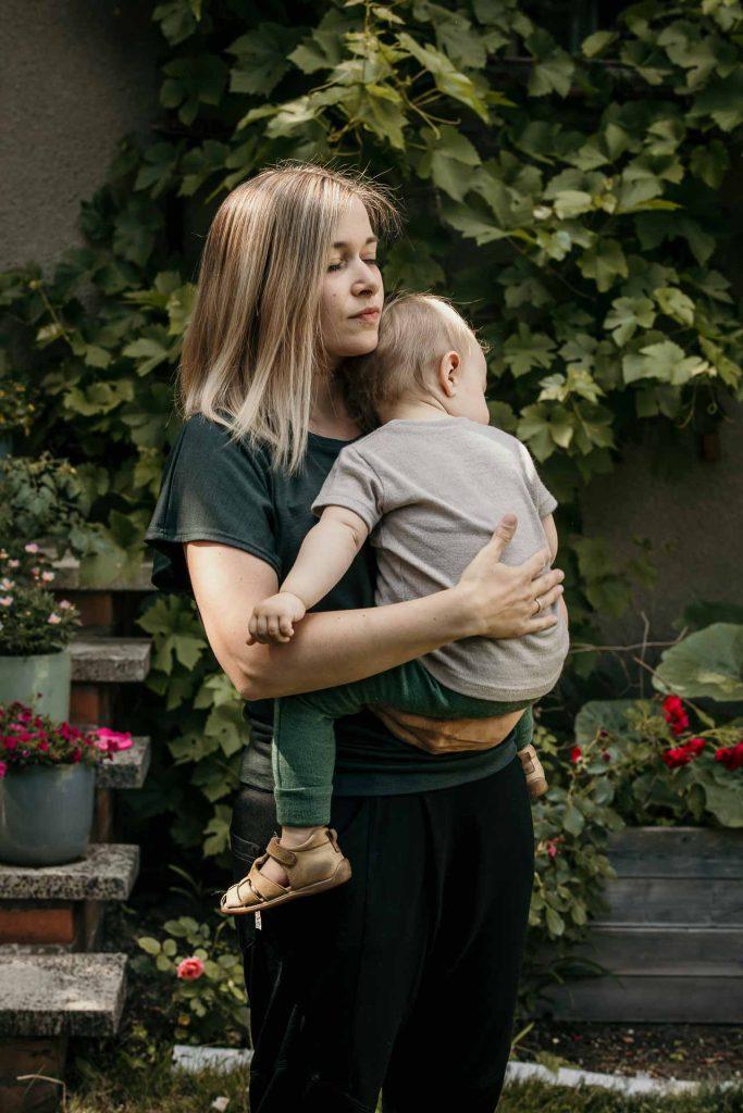 Mama steht im Garten mit kleiner Tochter auf dem Arm, beide kuscheln sich aneinander, Familienfotoshooting zu Hause Berlin